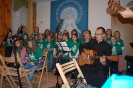 Świetlica Domu Nadziei, koncert Nutek Chwały ubranych w zielone koszulki z nazwą i logiem zespołu, w pierwszym planie z prawej stony ks. Tadeusz siedzi na krześle i gra na gitarze akustycznej, obok z lewej strony siedzą dwie dziewczynki grające na cymbałkach, w drugim planie dziewczęta śpiewają na stojąco, obok z lewej strony stoi dziewczynka grająca na skrzypcach, w tle duży obraz Matki Bożej